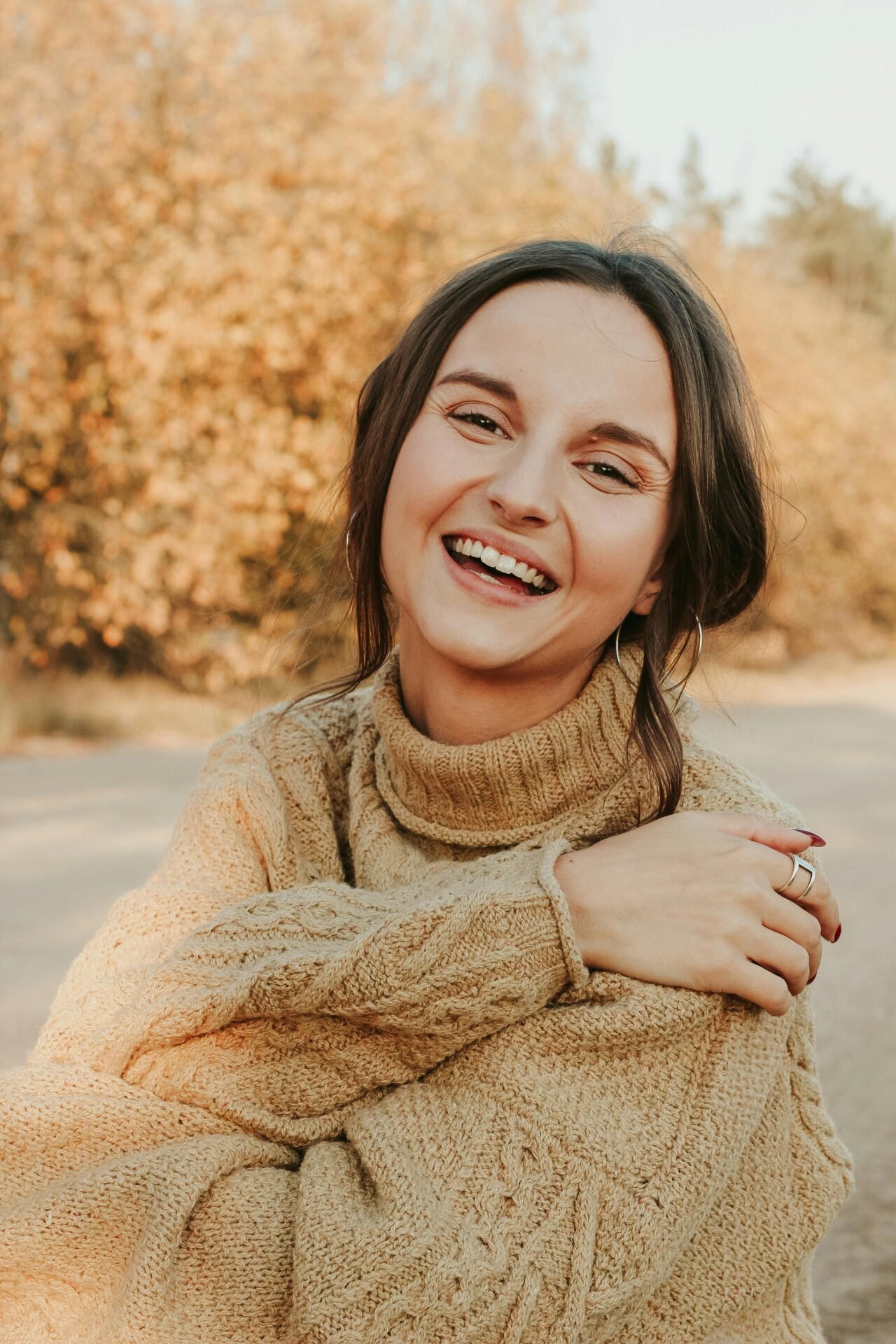 Cheerful woman smiling warmly in a cozy brown knitted sweater outdoors during fall.